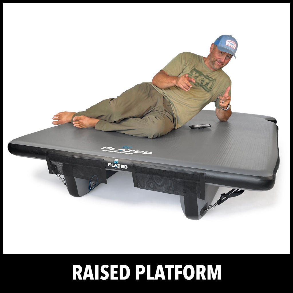 A man in casual clothing reclines on a large, elevated, black inflatable platform labeled FLATED. The platform is above the ground and supported by four sturdy inflatable legs. Text below reads RAISED PLATFORM.