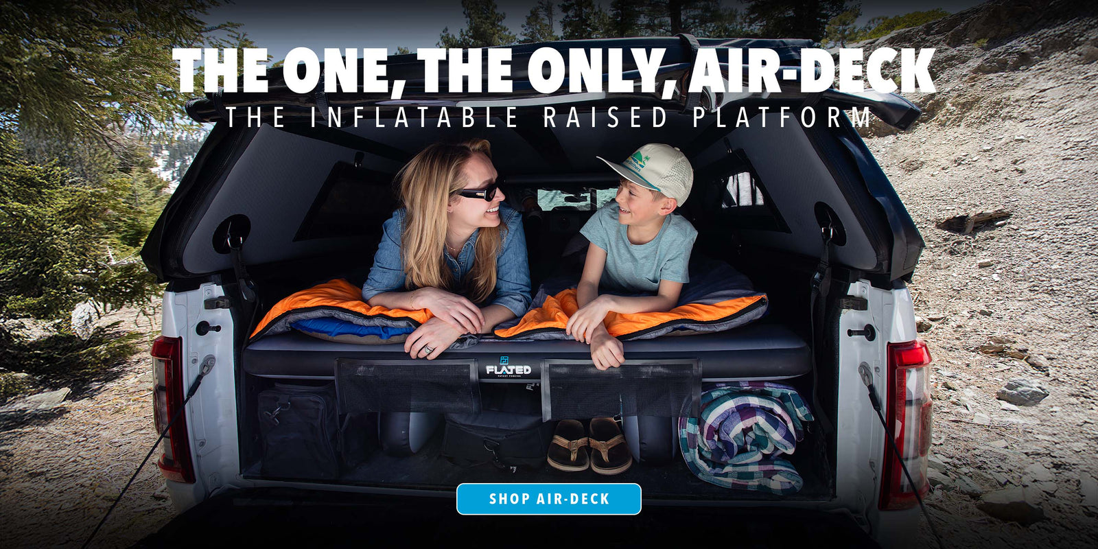 A woman and a boy lie inside the back of a truck fitted with an inflatable raised platform, smiling at each other. They are surrounded by sleeping bags and outdoor gear. Text promotes The One, The Only, Air-Deck.