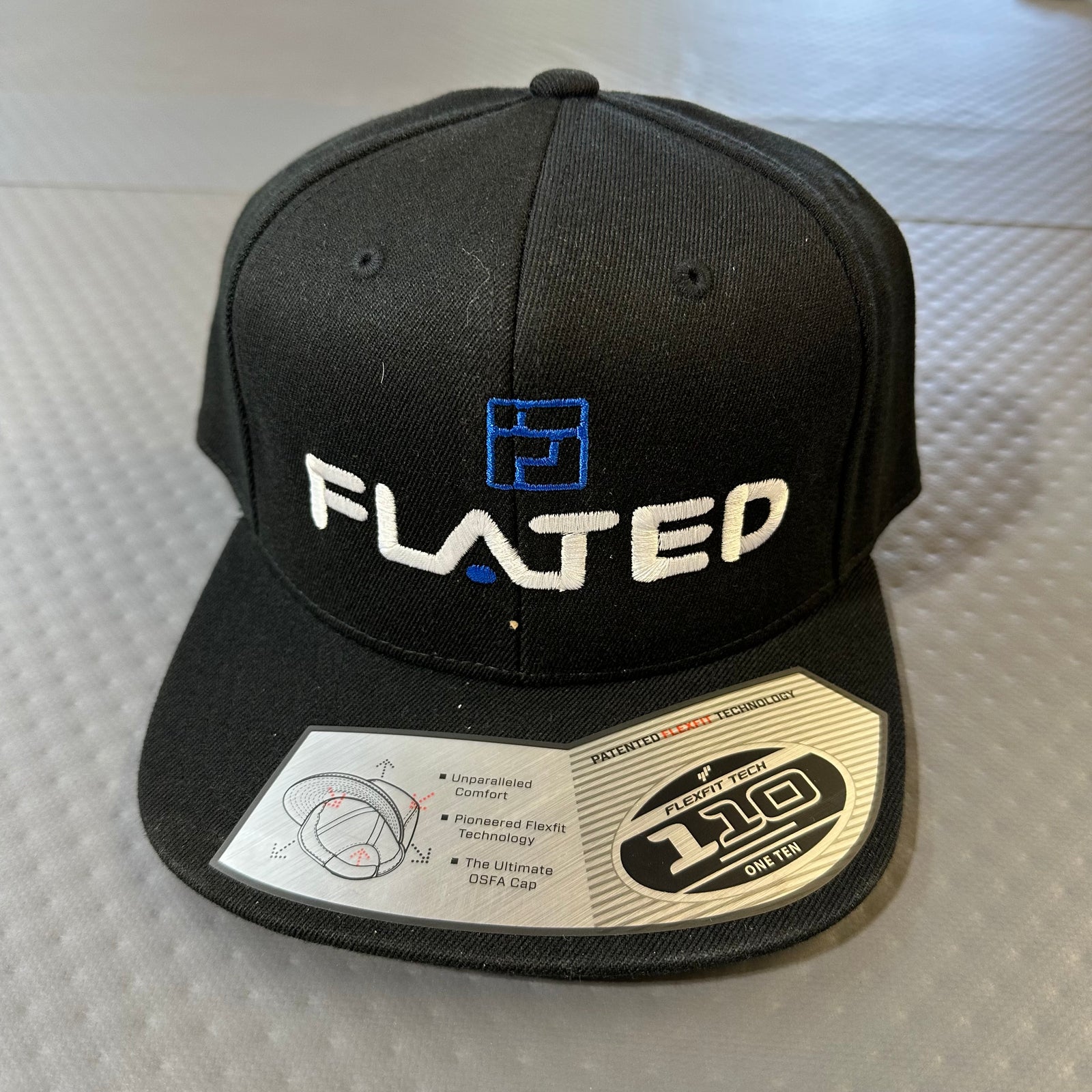The FLATED Team FLEXFIT Flat Brim Hat features a black design with white and blue FLATED embroidery, FLEXFIT 110 styling, and a silver sticker on the brim, shown on a textured gray surface.