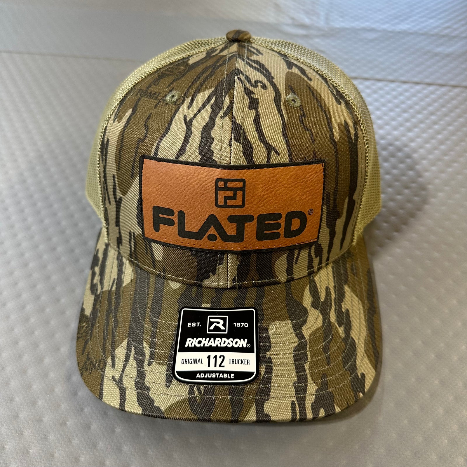 The FLATED Team Trucker Hat by FLATED features a black mesh-back design with "FLATED" embroidered in white and blue on the front, plus an adjustable sticker on the brim. The hat rests on a textured gray surface.