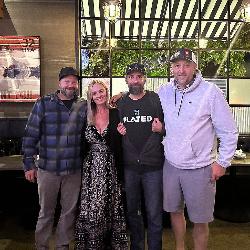 Four adults stand together inside a restaurant, smiling at the camera. Three men and one woman are posing, with a window and a striped awning in the background. The atmosphere looks casual and friendly.