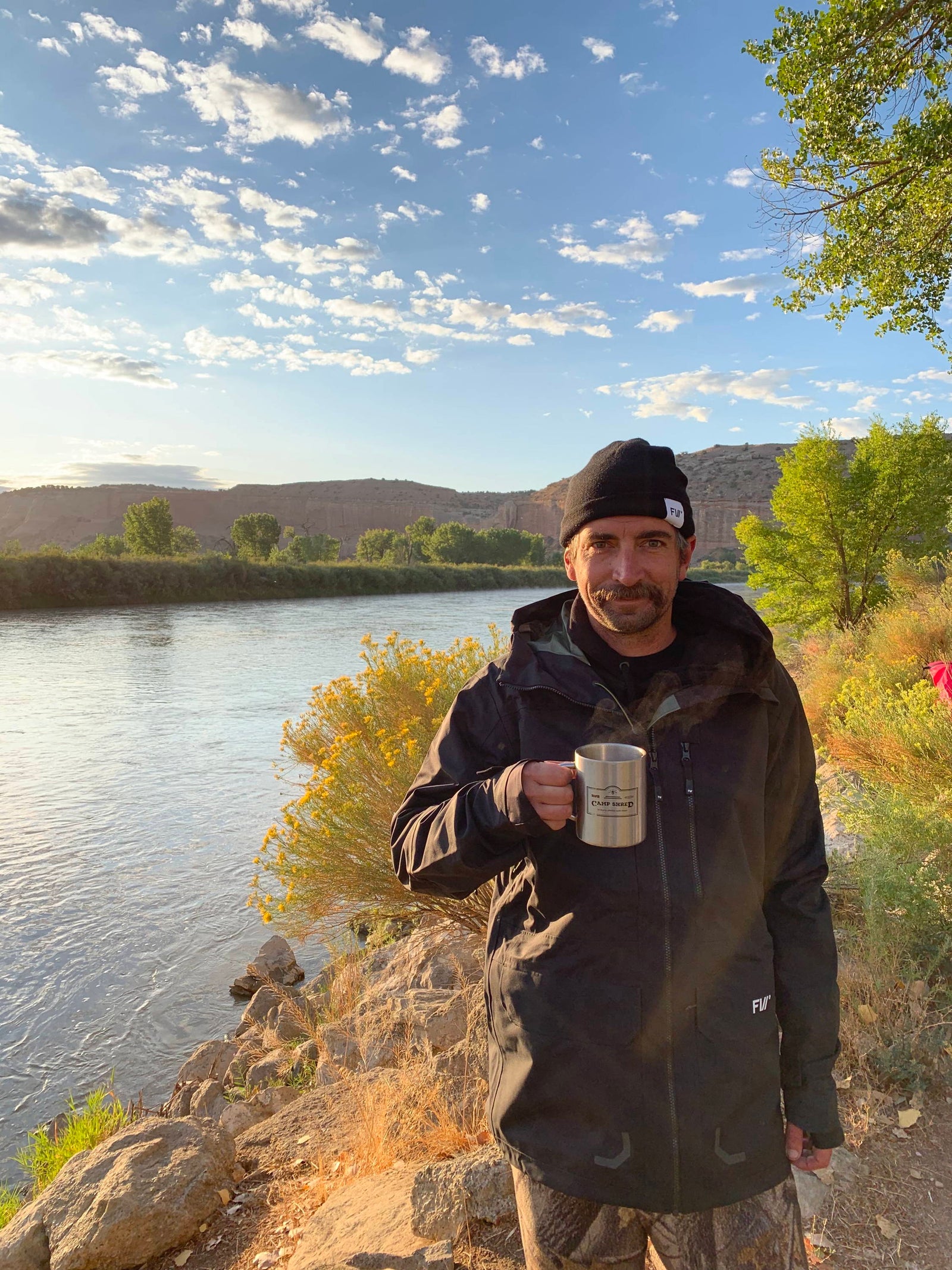 A man in a black jacket and beanie holds a metal mug while standing on rocky ground by a river, with trees and bushes nearby and hills in the background under a partly cloudy sky.
