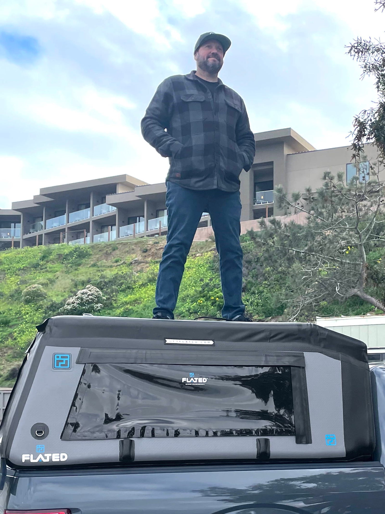 A man in a black cap and plaid jacket stands confidently on top of a gray rooftop tent mounted on a vehicle, with modern buildings and greenery in the background.