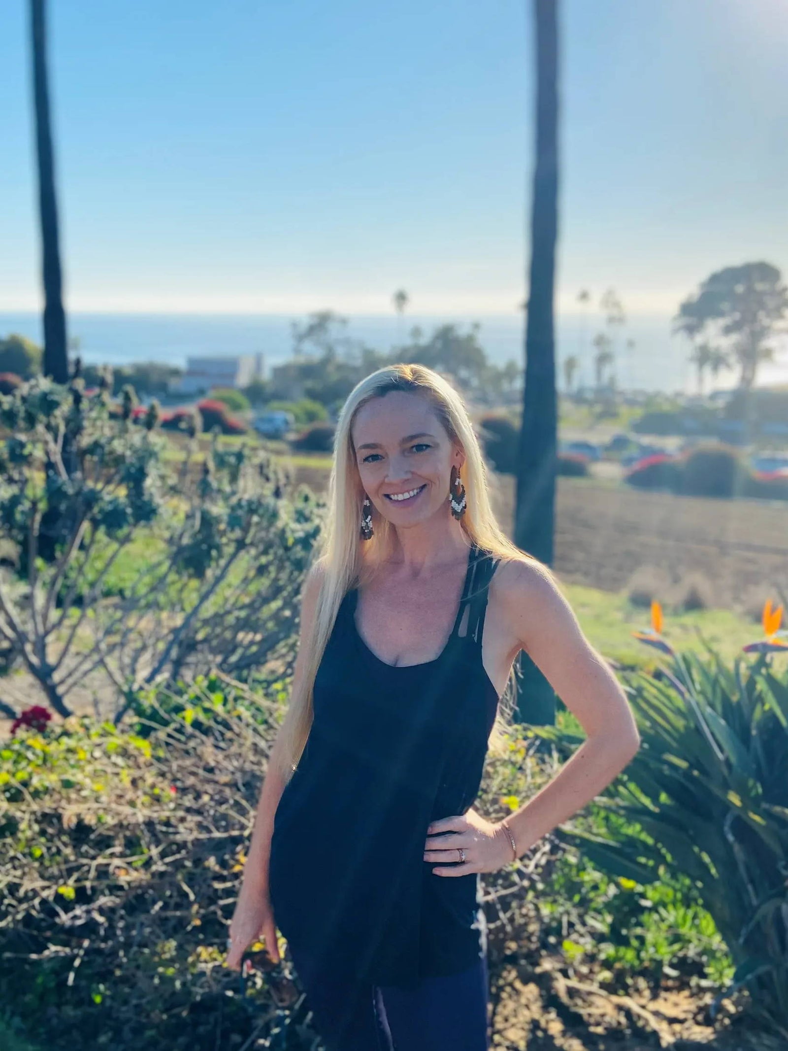 A woman with long blonde hair wearing a black sleeveless top stands outdoors, smiling with one hand on her hip. Behind her are plants, palm trees, cars, and a view of the ocean under a clear sky.
